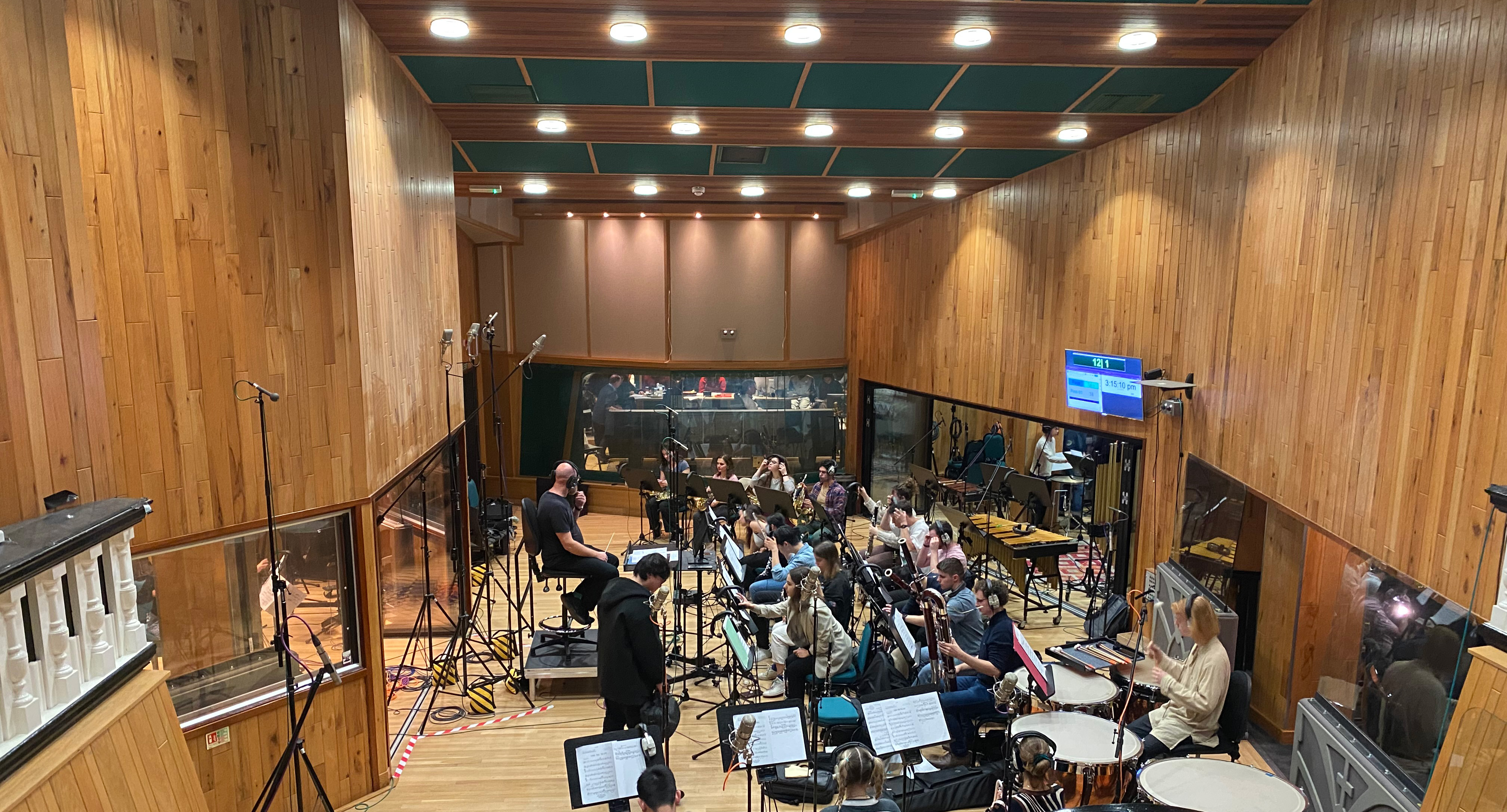 RCM Symphony Orchestra performing at Abbey Road Institute in a wood-panelled recording hall, with recording studio behind them.